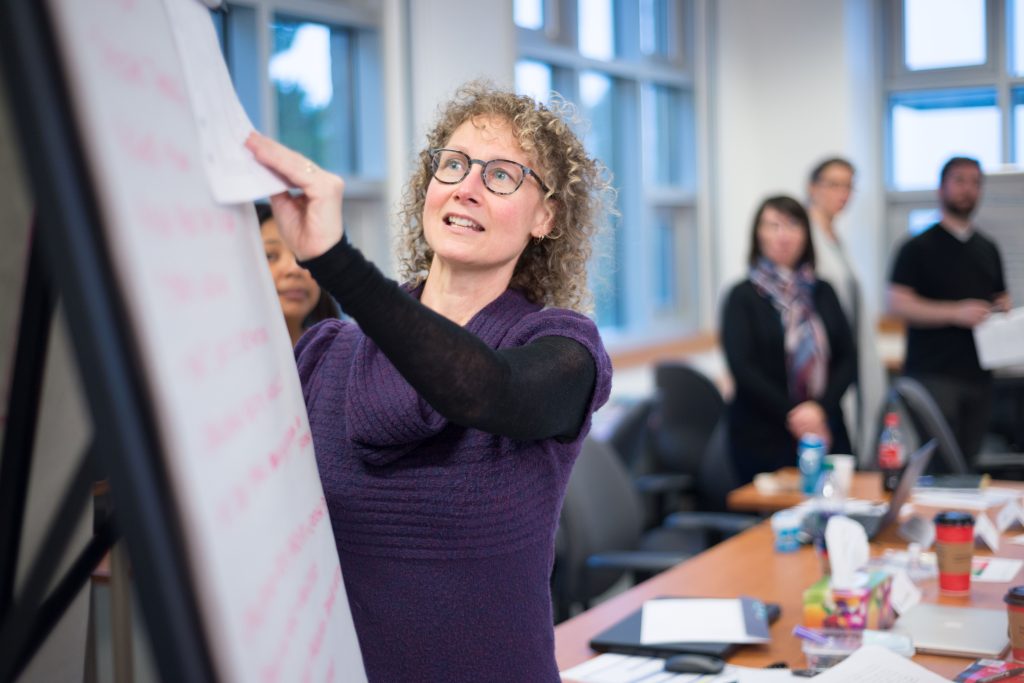 a facilitator leads a class, standing at a flipchart easel. Students look on from the background. 