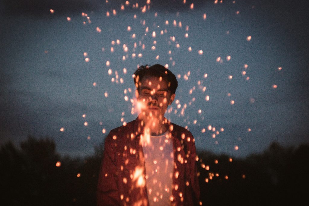 a person stands behind a campfire, with sparks shooting up in front of them, obscuring their view.